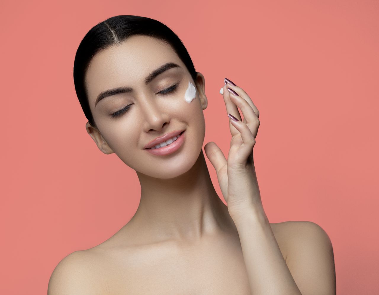 our-services-05 Studio portrait of a smiling woman applying cream on her face against a pink background.